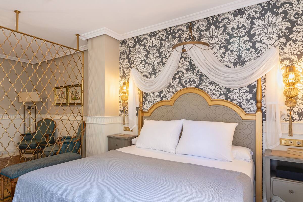 A four-poster bed with a canopy, a nightstand with a lamp, a chair, and a wall with a patterned wallpaper.