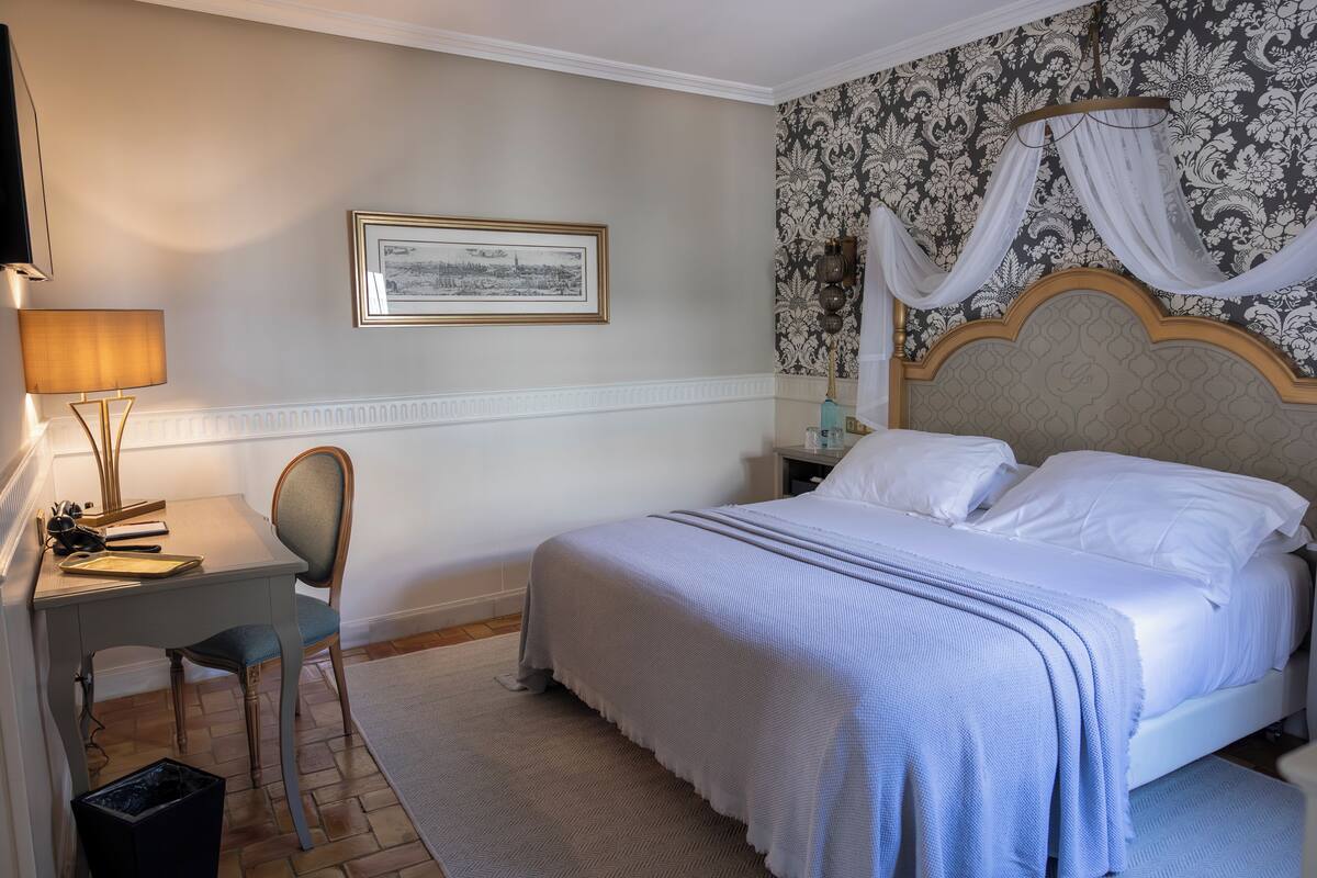A bedroom with a bed, a desk, a chair, a framed picture on the wall, and a patterned wallpaper.