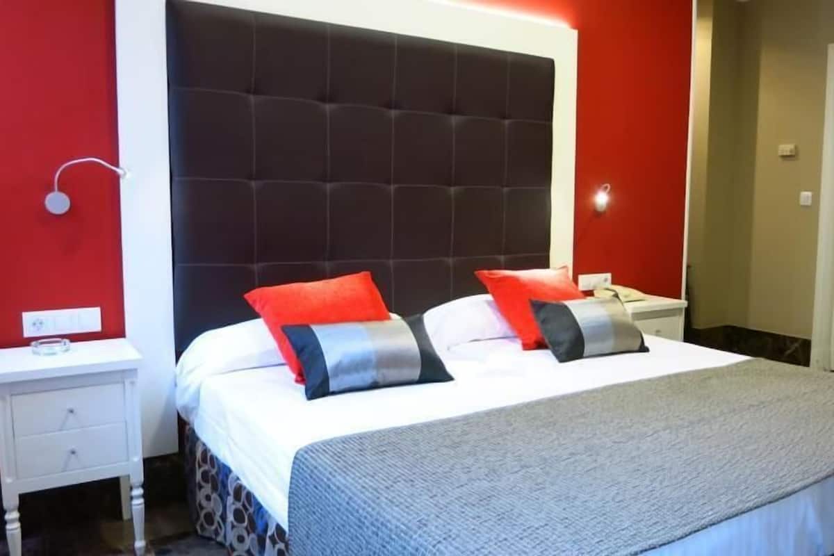 Habitación de hotel con una cama grande, dos mesitas de noche, una pared roja y una puerta.