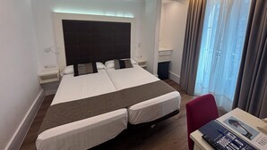 A hotel room with two beds, a desk, a chair, a window with curtains, and a ceiling light.