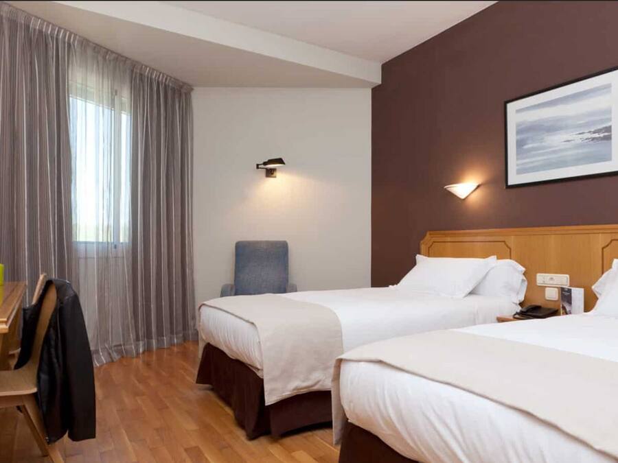 Habitación de hotel con dos camas, un escritorio con una computadora portátil, una silla y una ventana con cortinas.