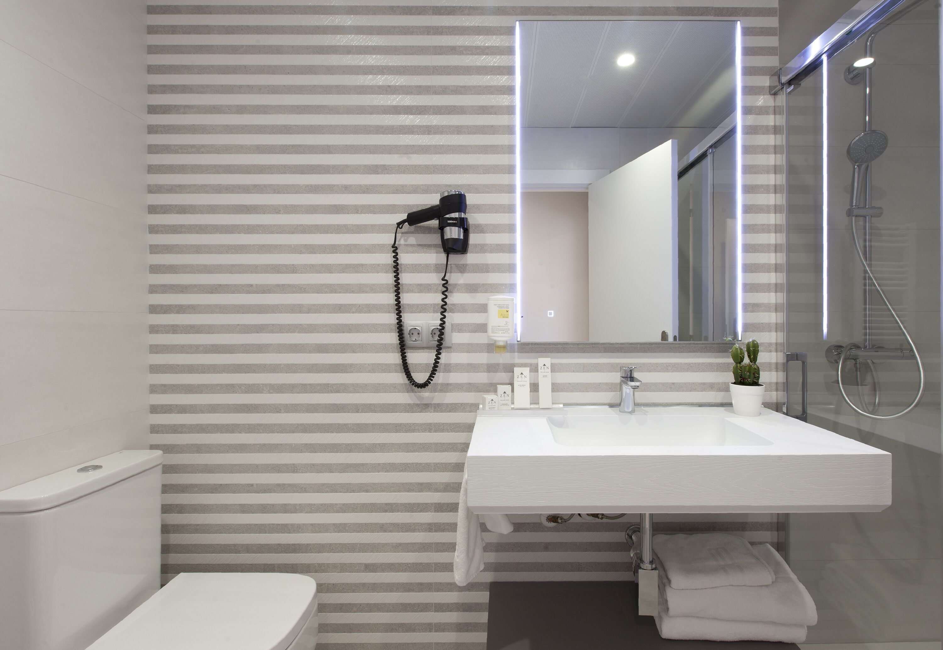 A modern bathroom with a striped wall, a white sink, a hairdryer, a shower, and a small potted plant.