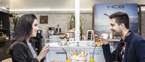 A breakfast setting with a table set for two, featuring a variety of breakfast items, a sign with the text "EAT WELL. FEEL WELL. LIVE WELL.", and a backdrop with a poster and a sign that reads "HCB".