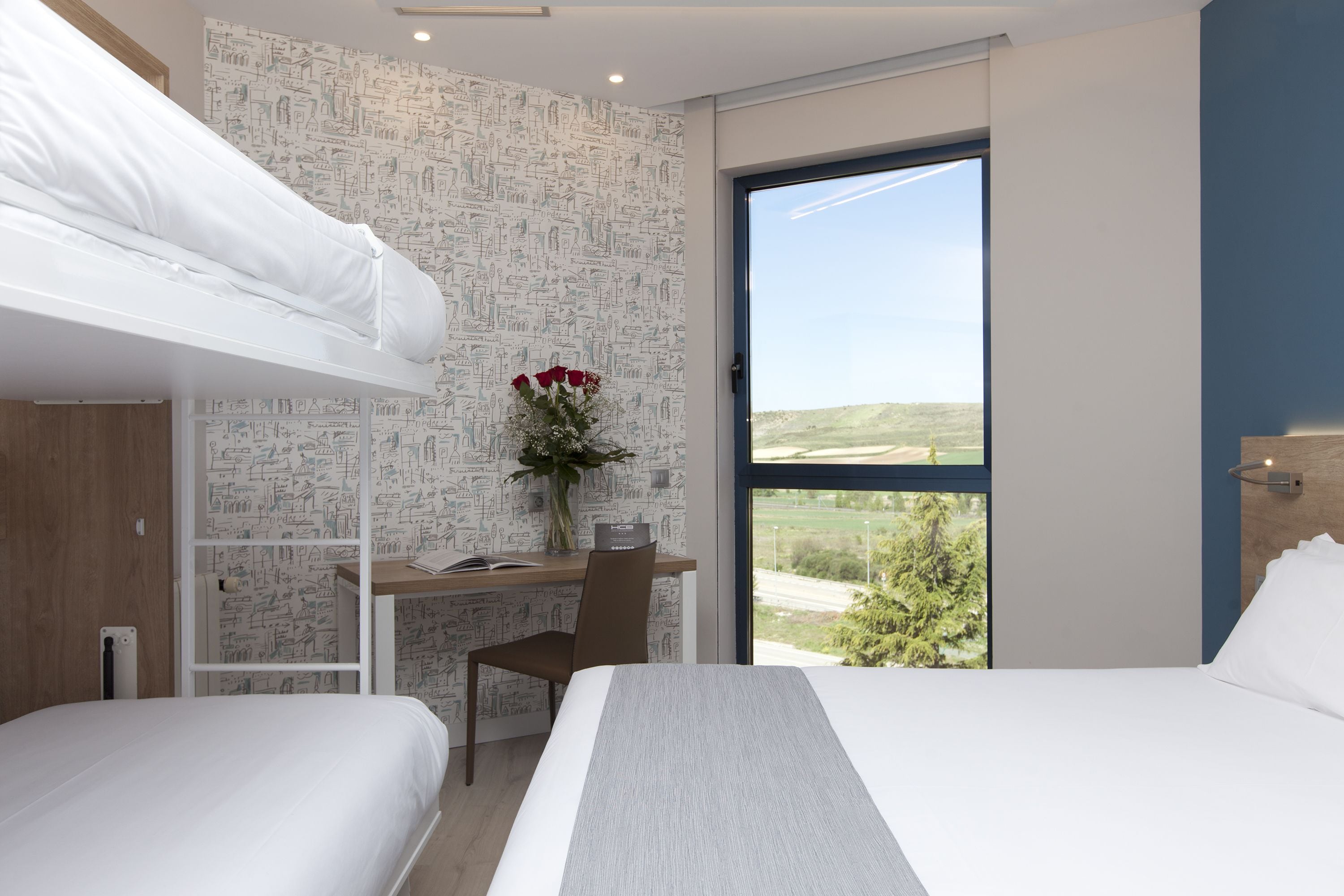 A modern hotel room with a bunk bed, a desk with a chair, a vase with flowers, and a large window offering a view of the outdoors.