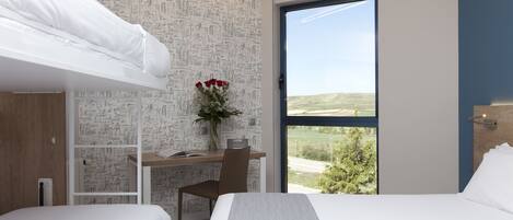 A modern hotel room with a bunk bed, a desk with a chair, a vase with flowers, and a large window offering a view of the outdoors.