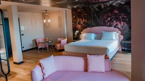 Una habitación de hotel moderna con una cama grande, un sofá rosa, una pequeña mesa redonda y un mural en el techo.