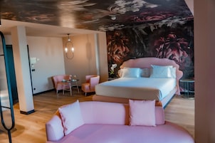 Una habitación de hotel moderna con una cama grande, un sofá rosa, una pequeña mesa redonda y un mural en el techo.