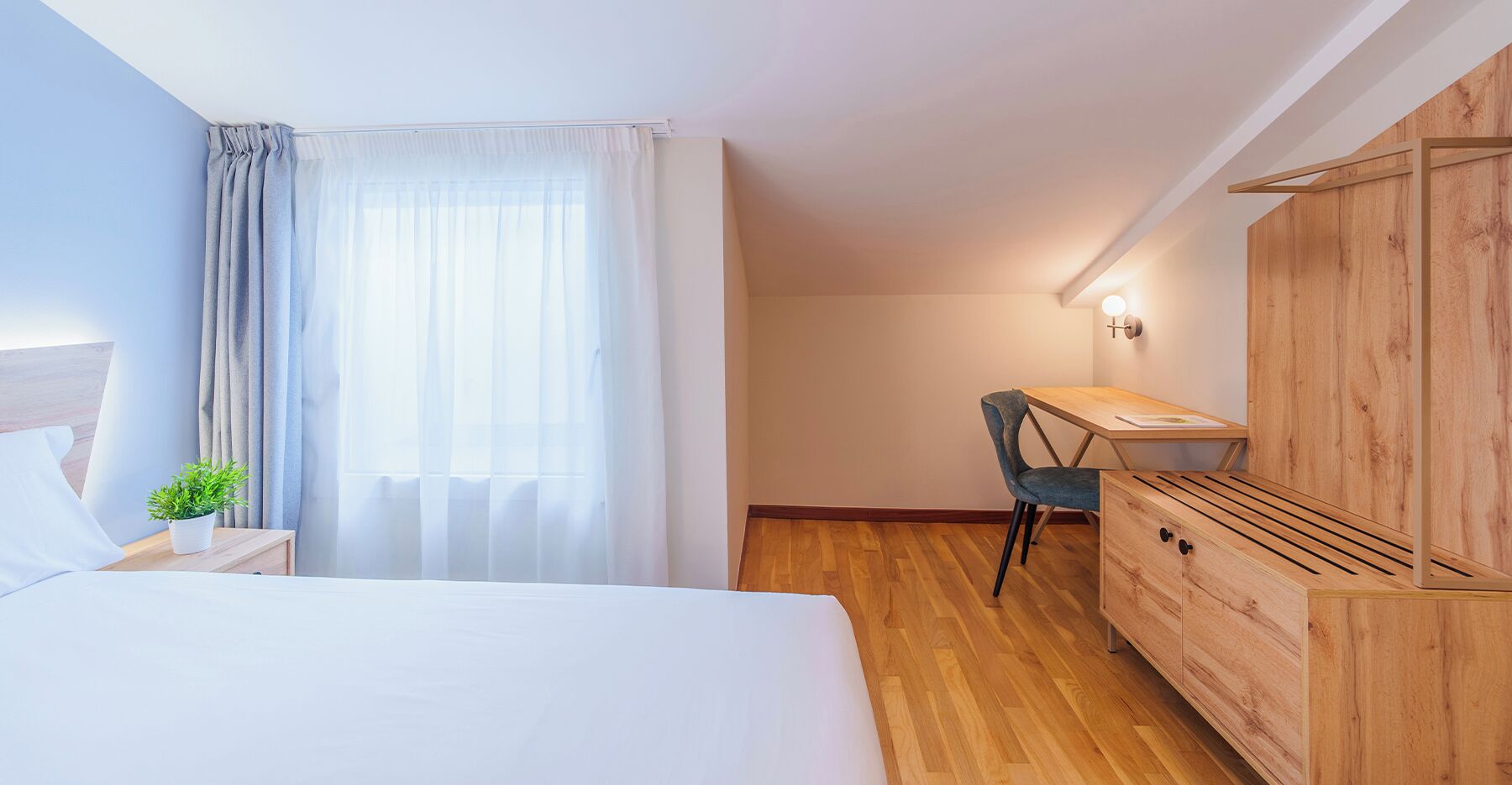 A bedroom with a bed, a desk, a chair, a wardrobe, and a window with curtains.
