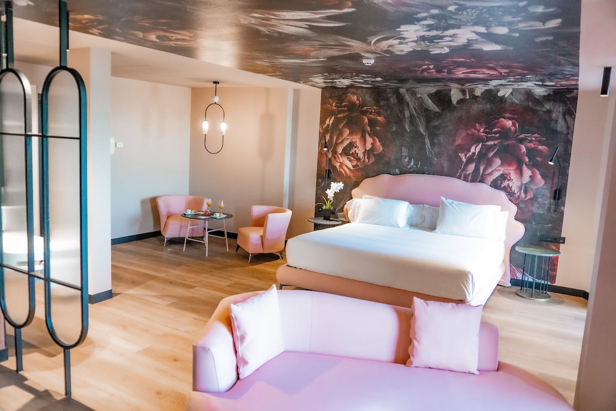 Un dormitorio moderno con una cama grande, dos sillones rosados, una mesita y un mural en el techo.