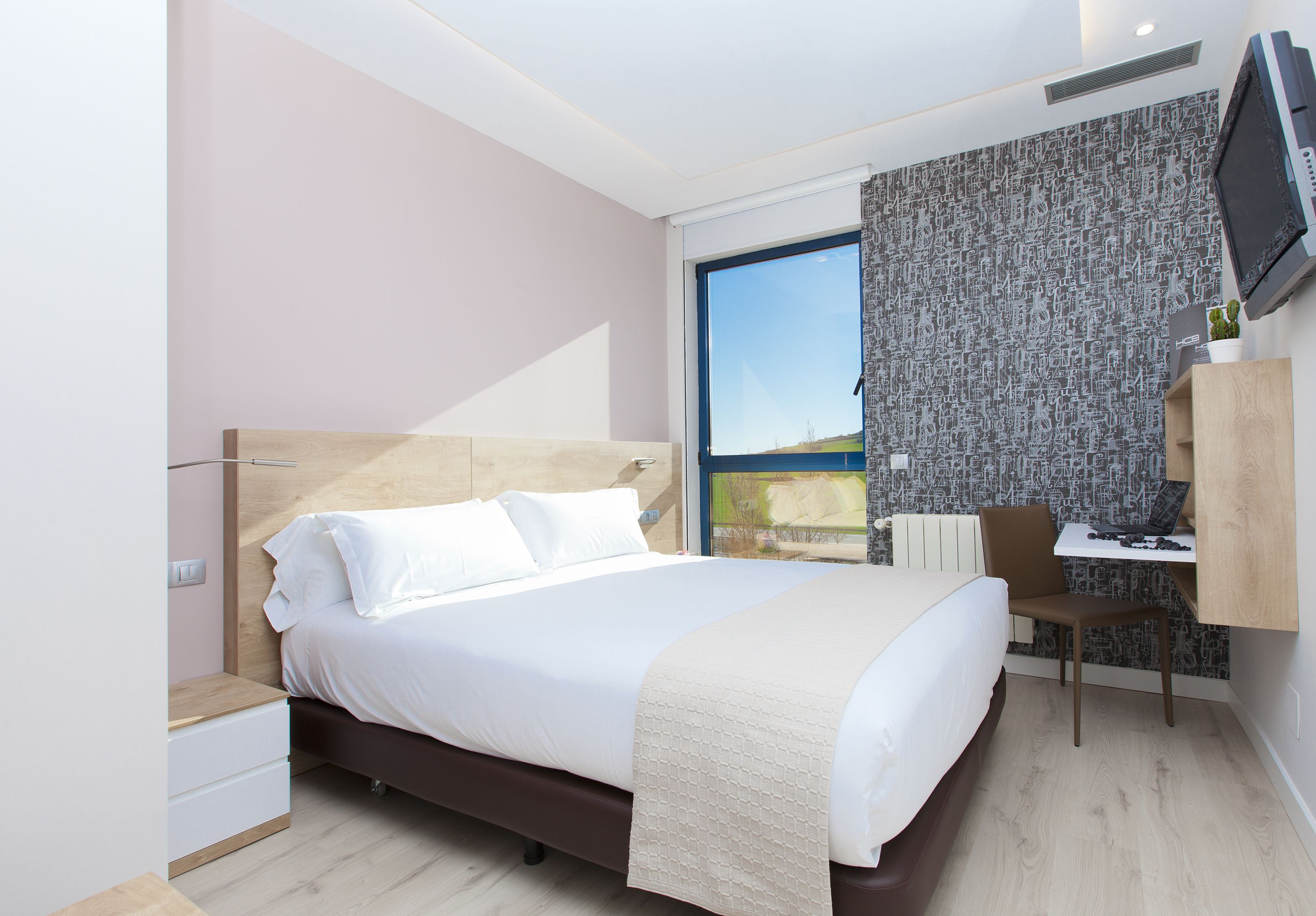 A modern hotel room with a large bed, a desk with a chair, a television, and a window with a view.