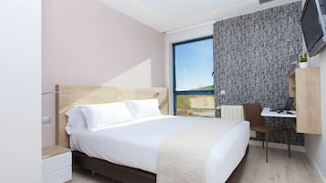 A modern hotel room with a large bed, a desk with a chair, a television, and a window with a view.