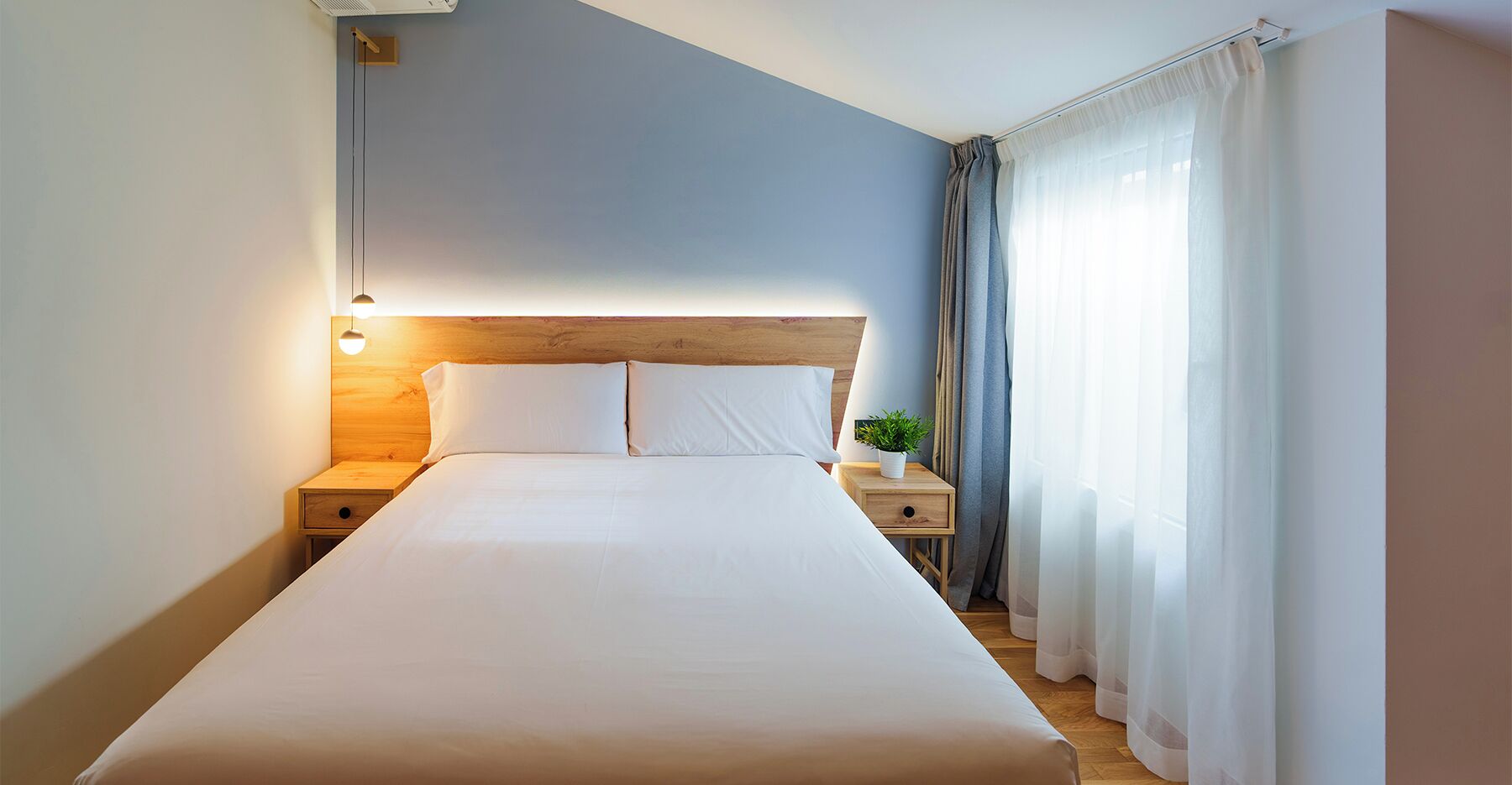 A neatly made bed in a hotel room with a wooden headboard, a bedside table with a lamp, and a potted plant.