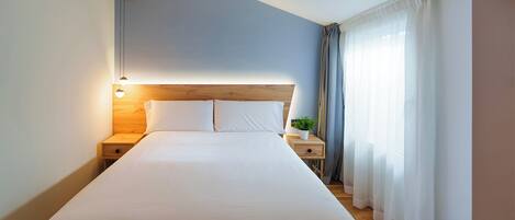 A neatly made bed in a hotel room with a wooden headboard, a bedside table with a lamp, and a potted plant.