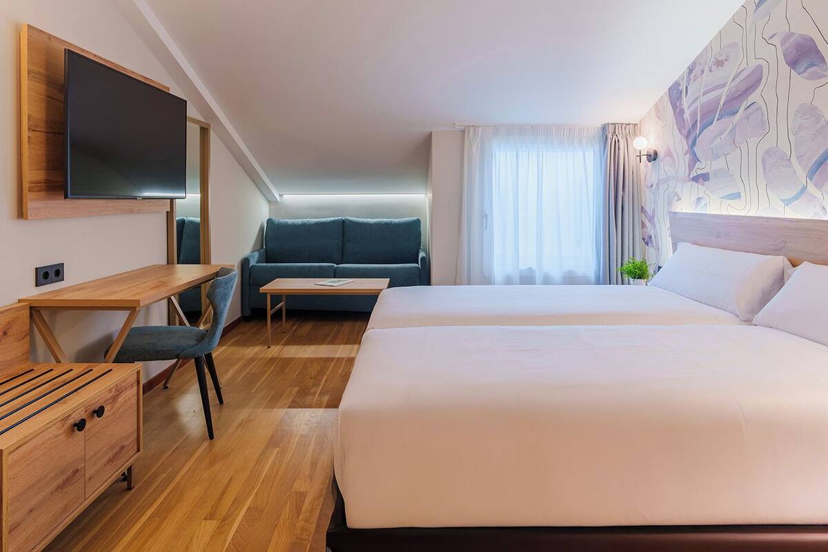 Habitación de hotel con una cama grande, un escritorio con silla, un sofá y un televisor instalado en la pared.