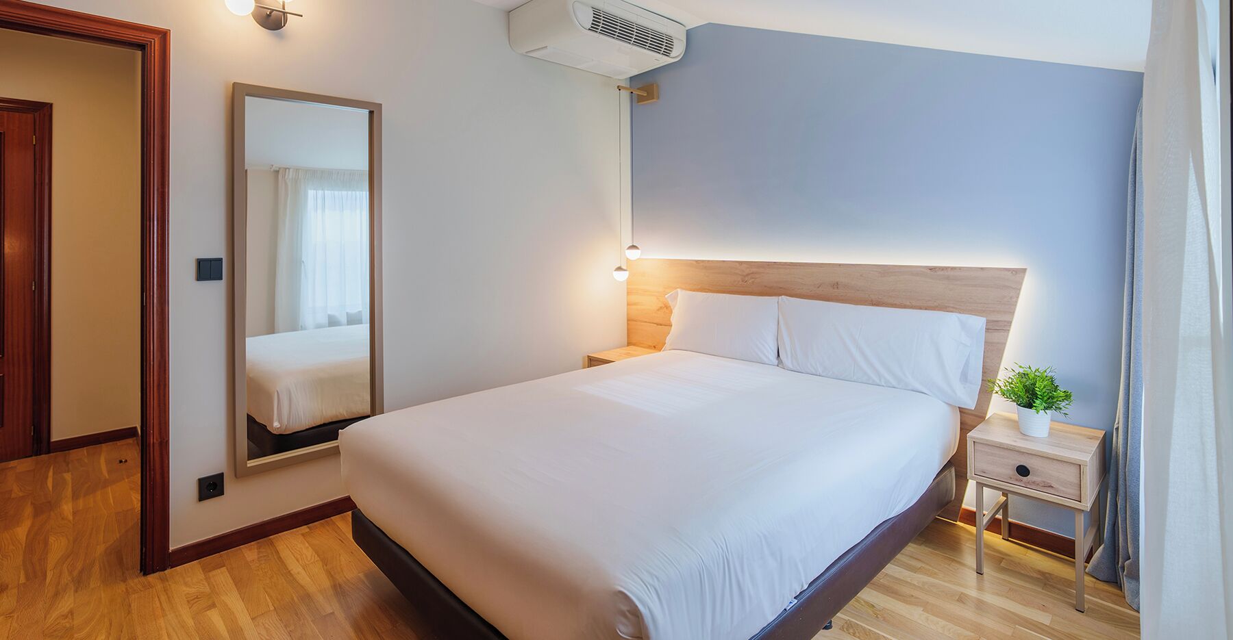 Une chambre d’hôtel avec un grand lit, une table de chevet avec une lampe, un miroir et une petite plante.