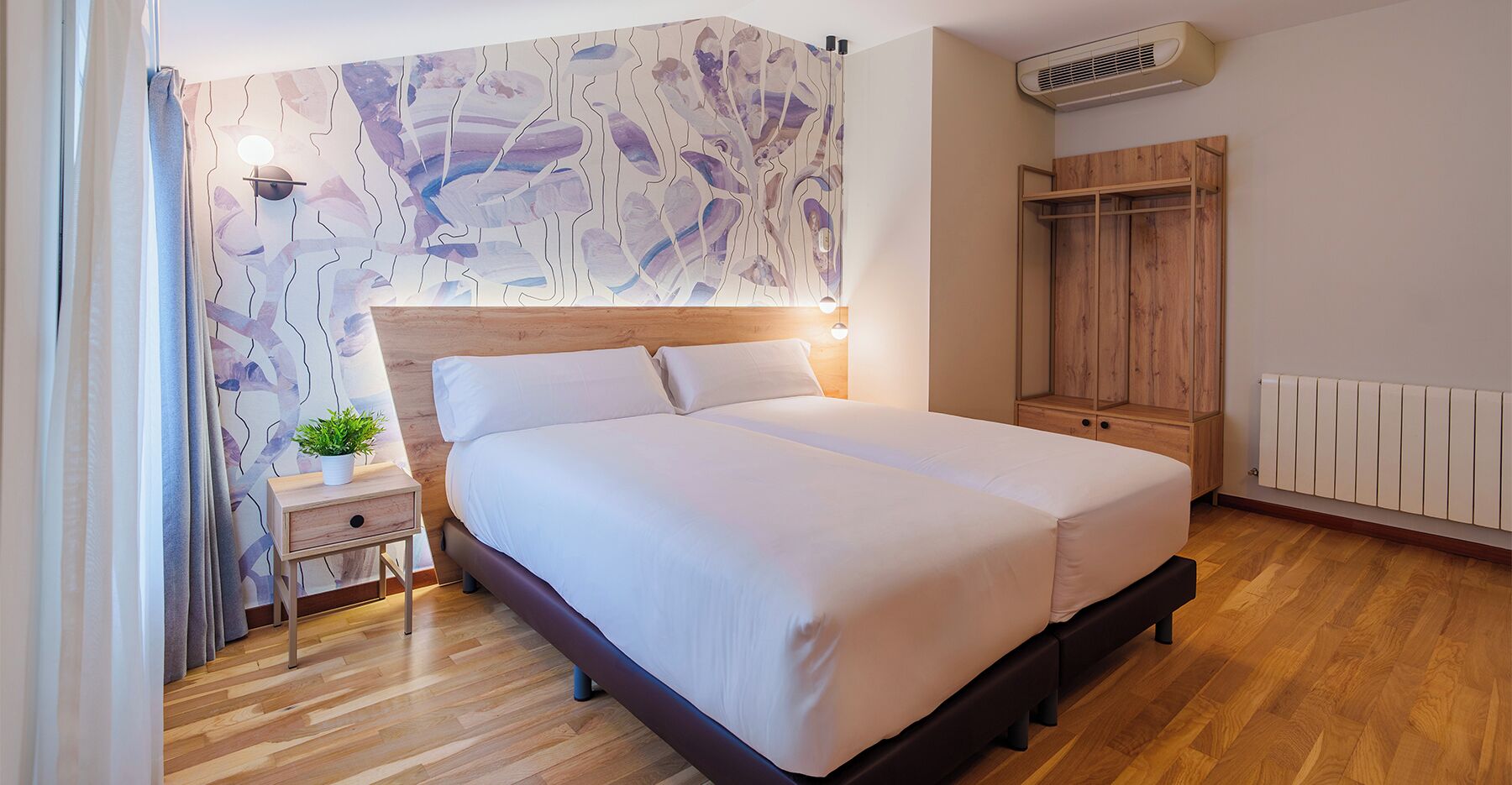 Un dormitorio moderno con cabecera de madera, cama grande, mesita de noche con una planta, armario y radiador.