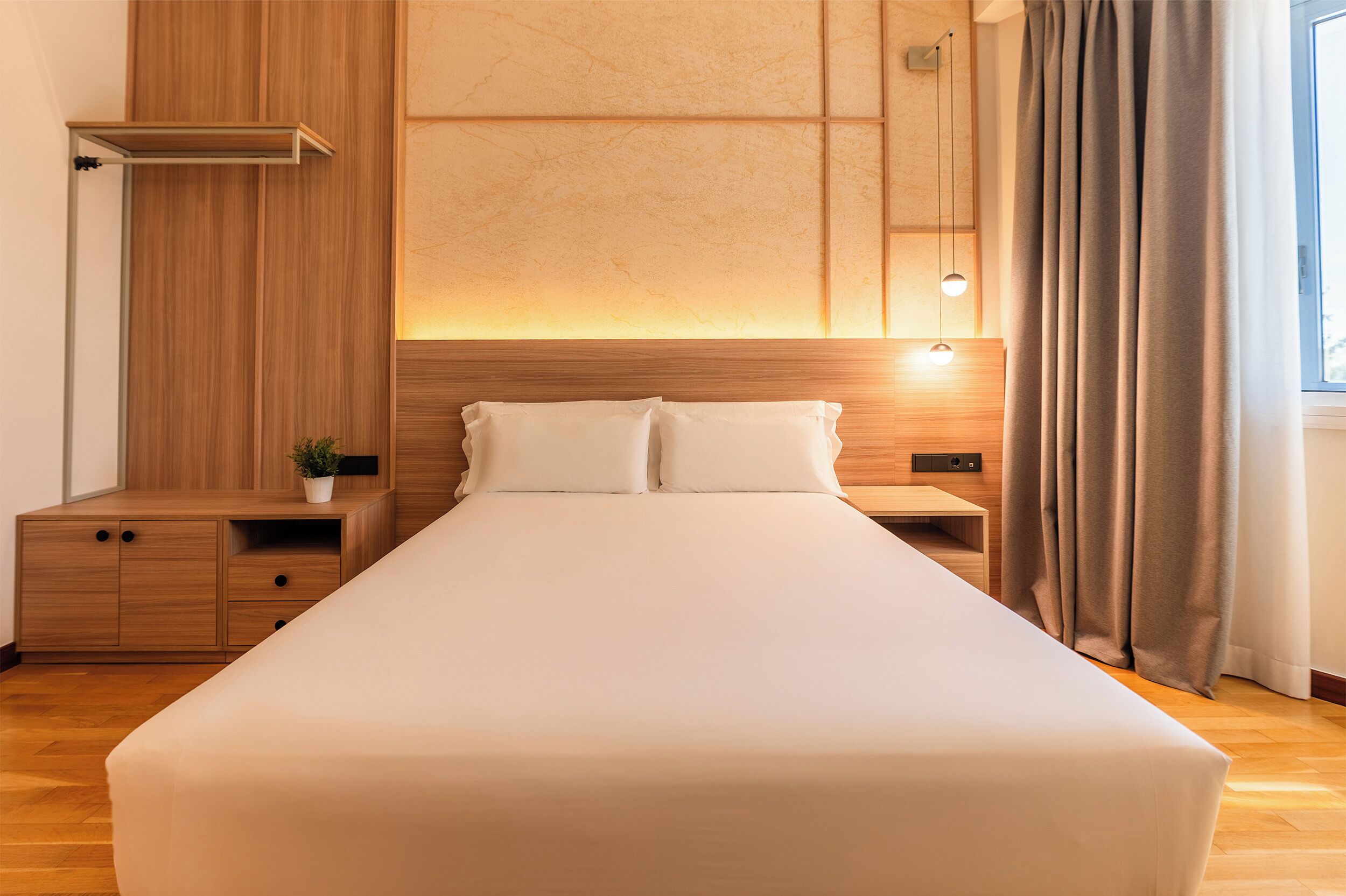 A hotel room with a large bed, wooden nightstands, a bedside table, a wall-mounted shelf, and a window with curtains.