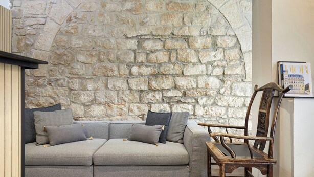 A room with a stone wall, a grey sofa with cushions, and a wooden chair. - Hotel Cortes Rambla (Barcelona)
