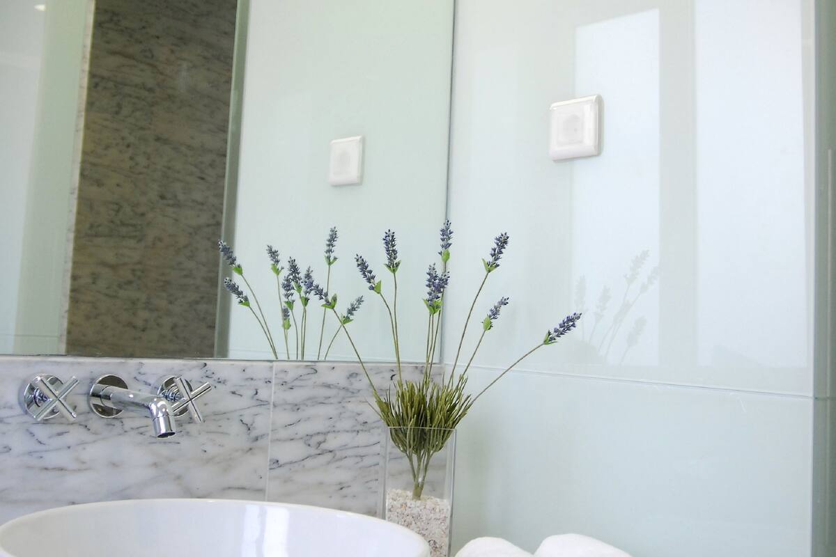 Un baño con un lavamanos grande de color blanco, encimera de mármol, cabina de ducha con paredes de vidrio y un jarrón con flores de lavanda.