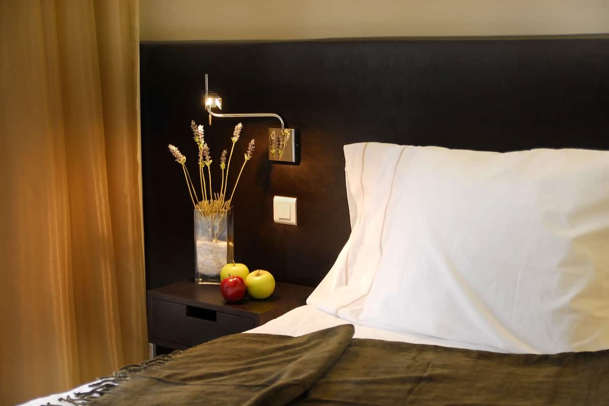 Una cama bien hecha con almohadas blancas y un cabecero oscuro. Una mesita de noche al lado de la cama sostiene un jarrón con flores secas y dos manzanas.