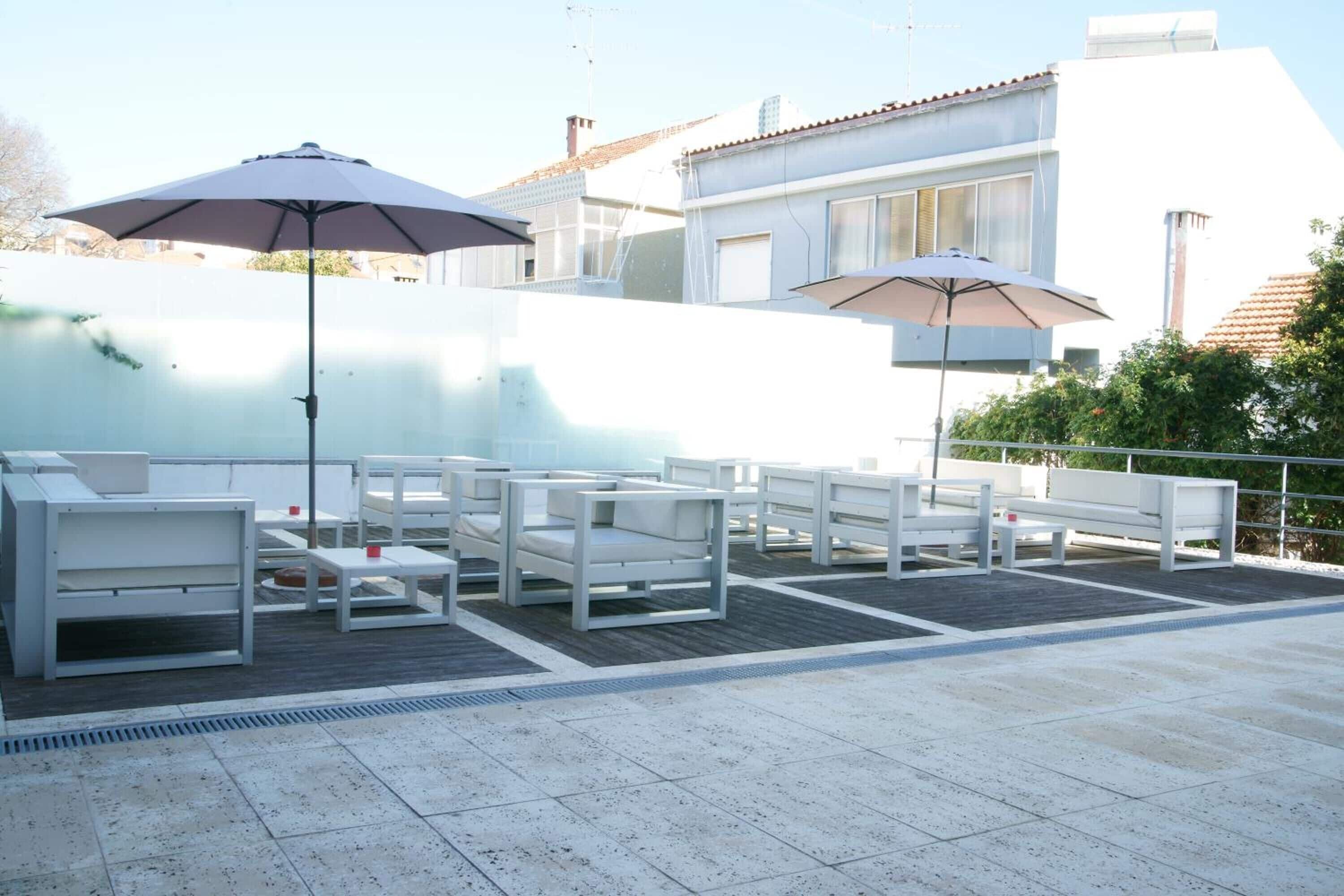 Un área de estar al aire libre con mobiliario blanco y sombrillas, piso de baldosas y un edificio blanco al fondo.