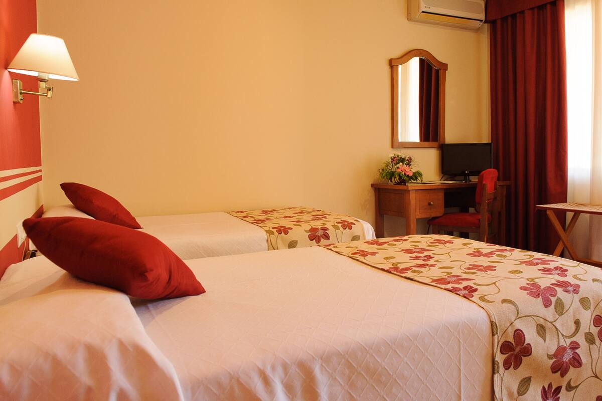 A hotel room with two beds, a desk, a chair, a mirror, and a window with curtains.