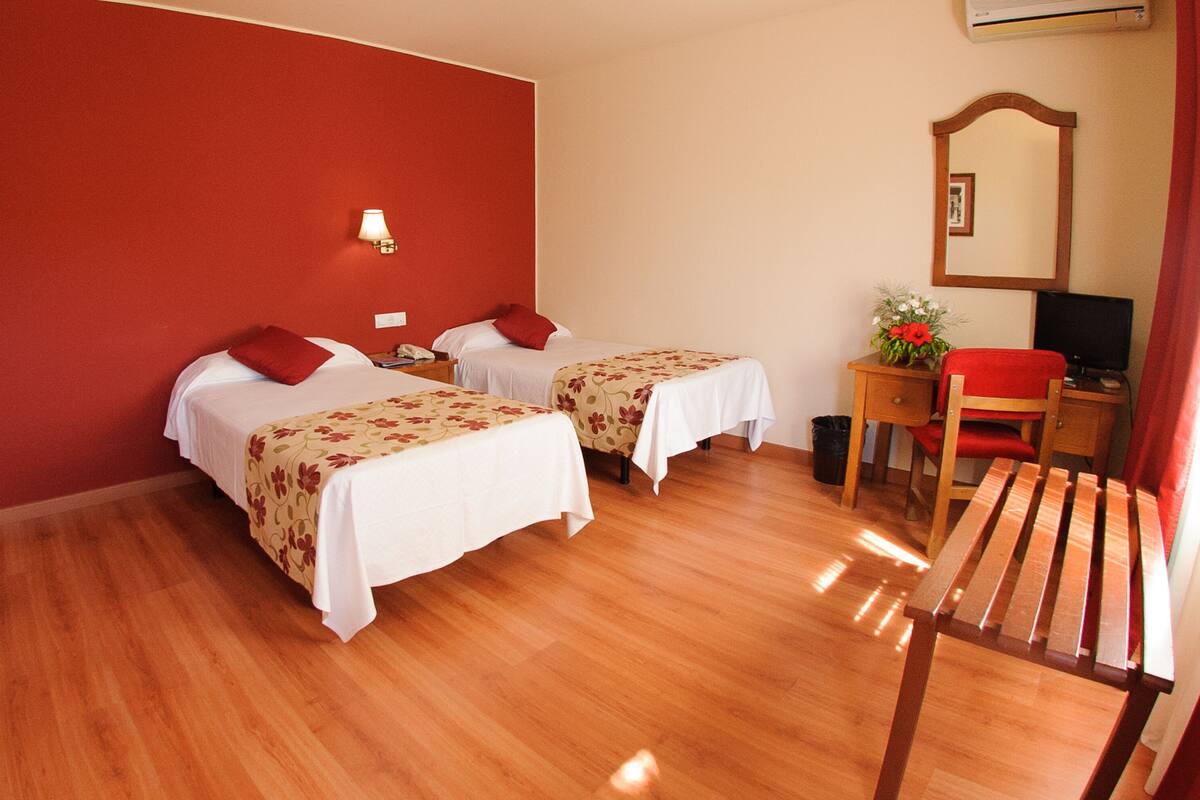 A hotel room with two beds, a desk with a chair, a mirror, and a small trash bin.