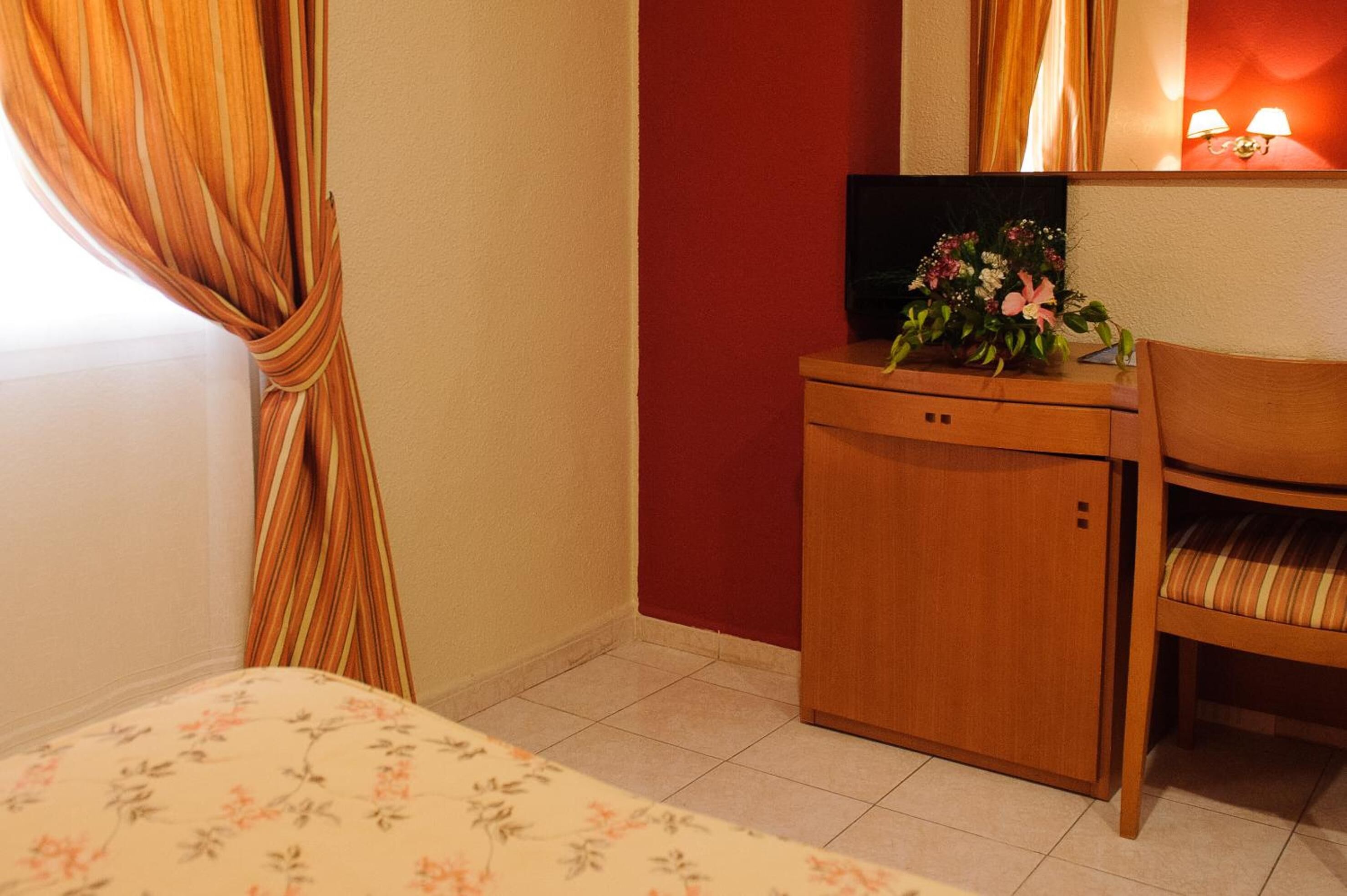 Habitación de hotel con cama, un escritorio de madera con televisor y flores, una silla y pared roja.