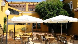 Une terrasse extérieure avec des tables et des chaises, des parasols blancs et un bâtiment jaune avec un toit en tuiles.