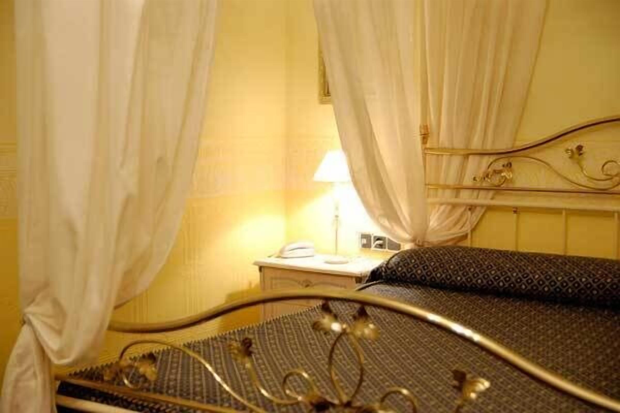 Una cama con estructura metálica y una colcha estampada, una mesita de noche con una lámpara y cortinas blancas.