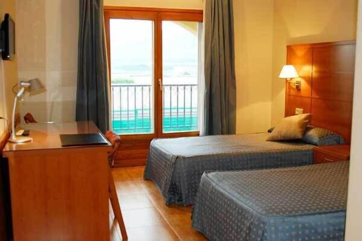 A hotel room with two single beds, a desk with a lamp, a wooden headboard, and a view through a sliding door.