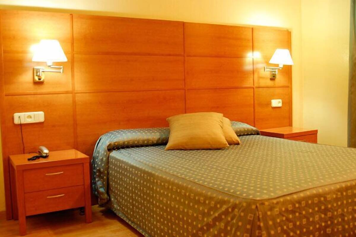 A hotel room with a wooden headboard, two bedside lamps, a bed with a patterned bedspread, and a nightstand with a phone and a small drawer.