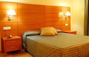 Habitación de hotel con cabecera de madera, dos lámparas de noche, una cama con colcha estampada y una mesita de noche con teléfono y un cajón pequeño.