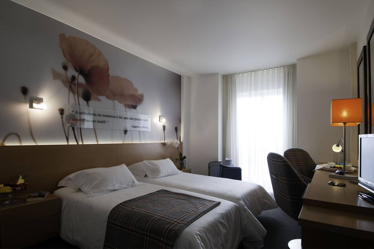 A hotel room with two beds, a desk with a lamp, a chair, and a wall with a quote and flowers.