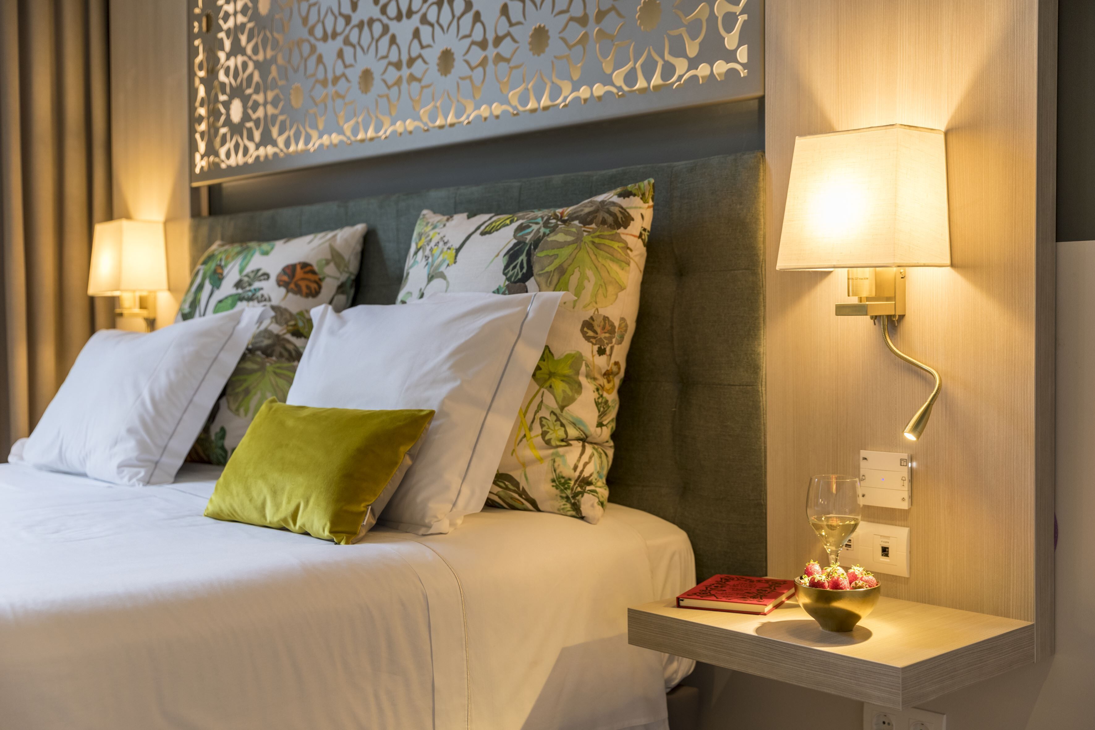Un lit bien fait, avec des oreillers blancs et verts, une table de chevet sur laquelle se trouvent un livre et un verre de vin, et une lampe fixée au mur.