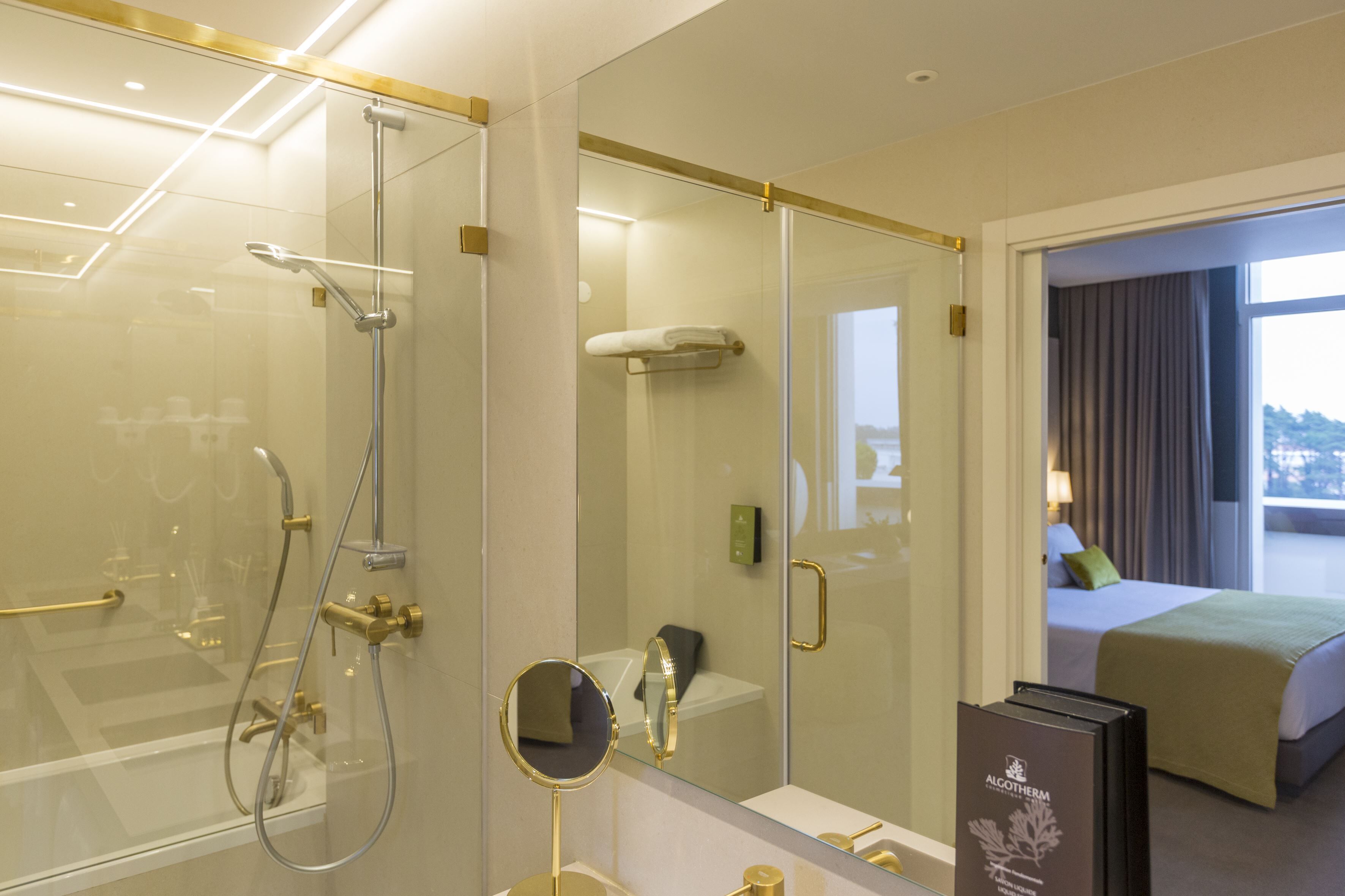 A hotel room with a glass-enclosed shower, a bedside table with a mirror, and a bed with a green blanket.