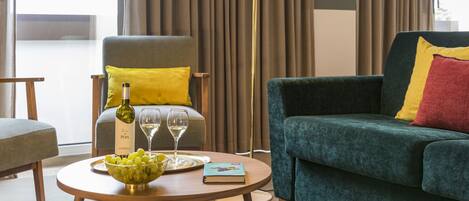 Una sala de estar moderna con un sofá verde, una almohada amarilla, una mesa de centro de madera con un cuenco de uvas y copas de vino, y una lámpara de pie.