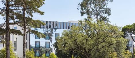 Un edificio moderno con amplios ventanales y techo plano, rodeado de árboles y un área pavimentada.