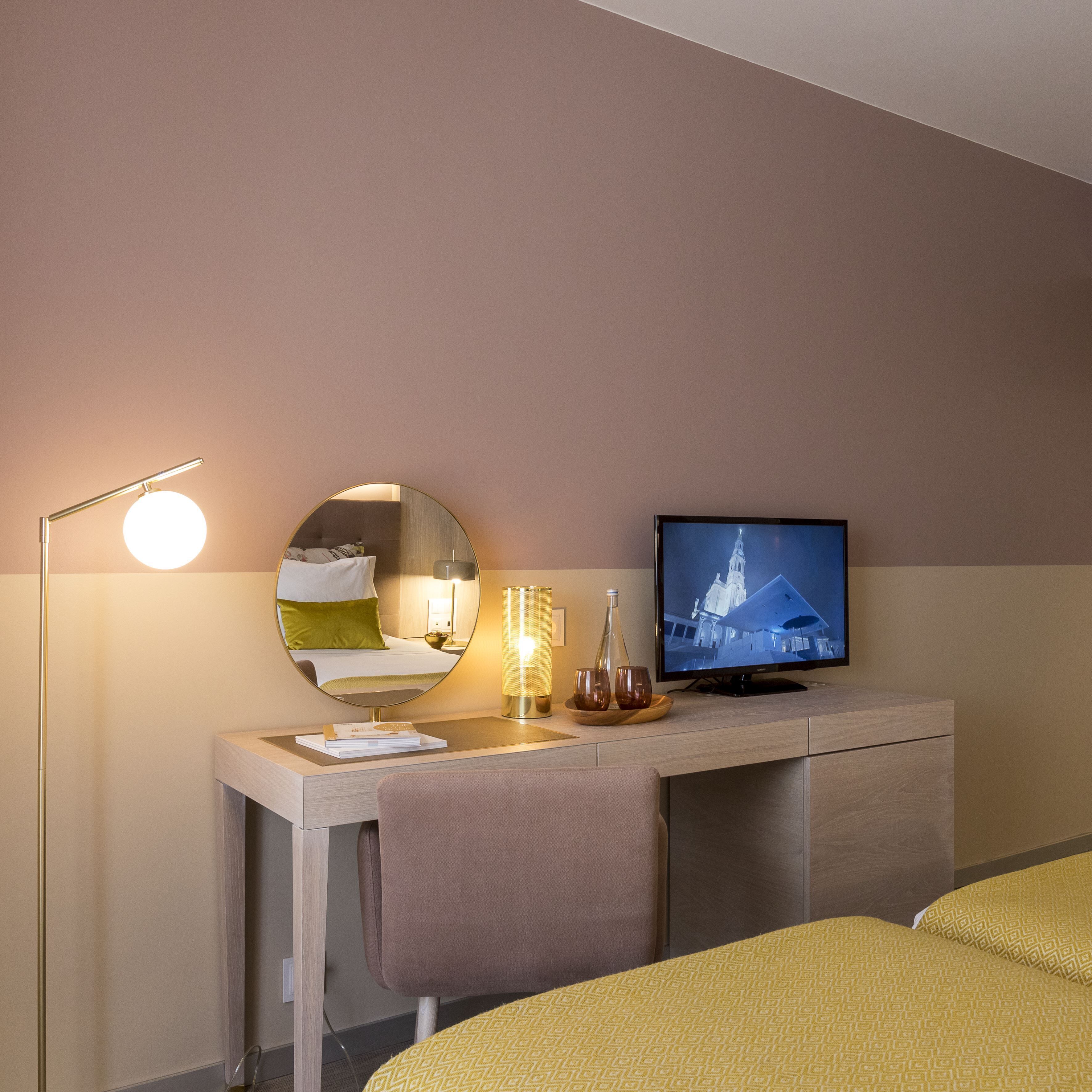 Habitación de hotel con cama, escritorio con televisor de pantalla plana, espejo redondo, lámpara de pie y mesita de noche con lámpara y vasos.
