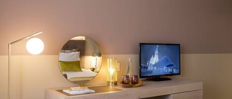 Habitación de hotel con cama, escritorio con televisor de pantalla plana, espejo redondo, lámpara de pie y mesita de noche con lámpara y vasos.
