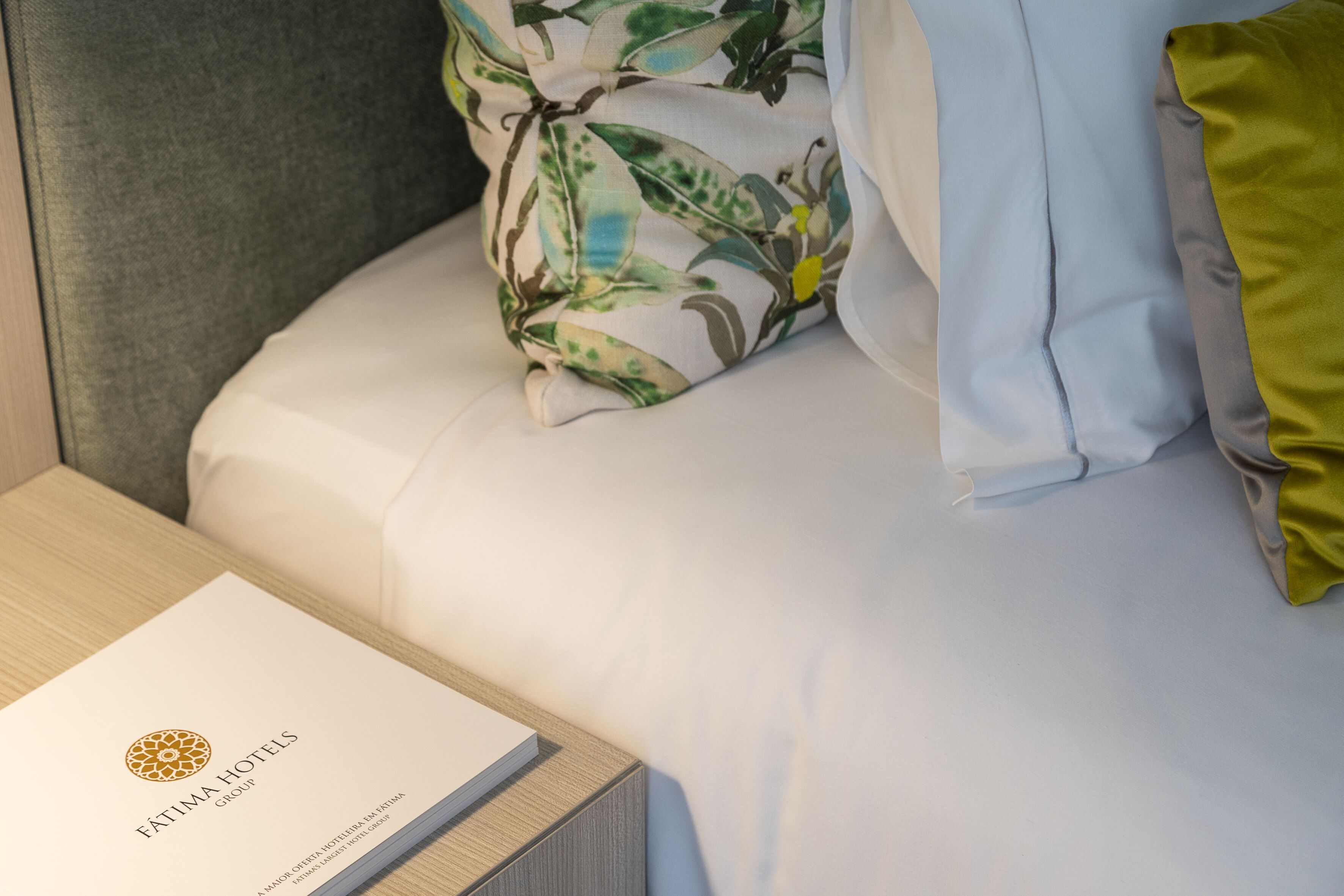 Una cama bien hecha con ropa blanca y una almohada floral, una mesita de noche de madera con un libro titulado "FATIMA HOTELS Group" y un cabecero gris.