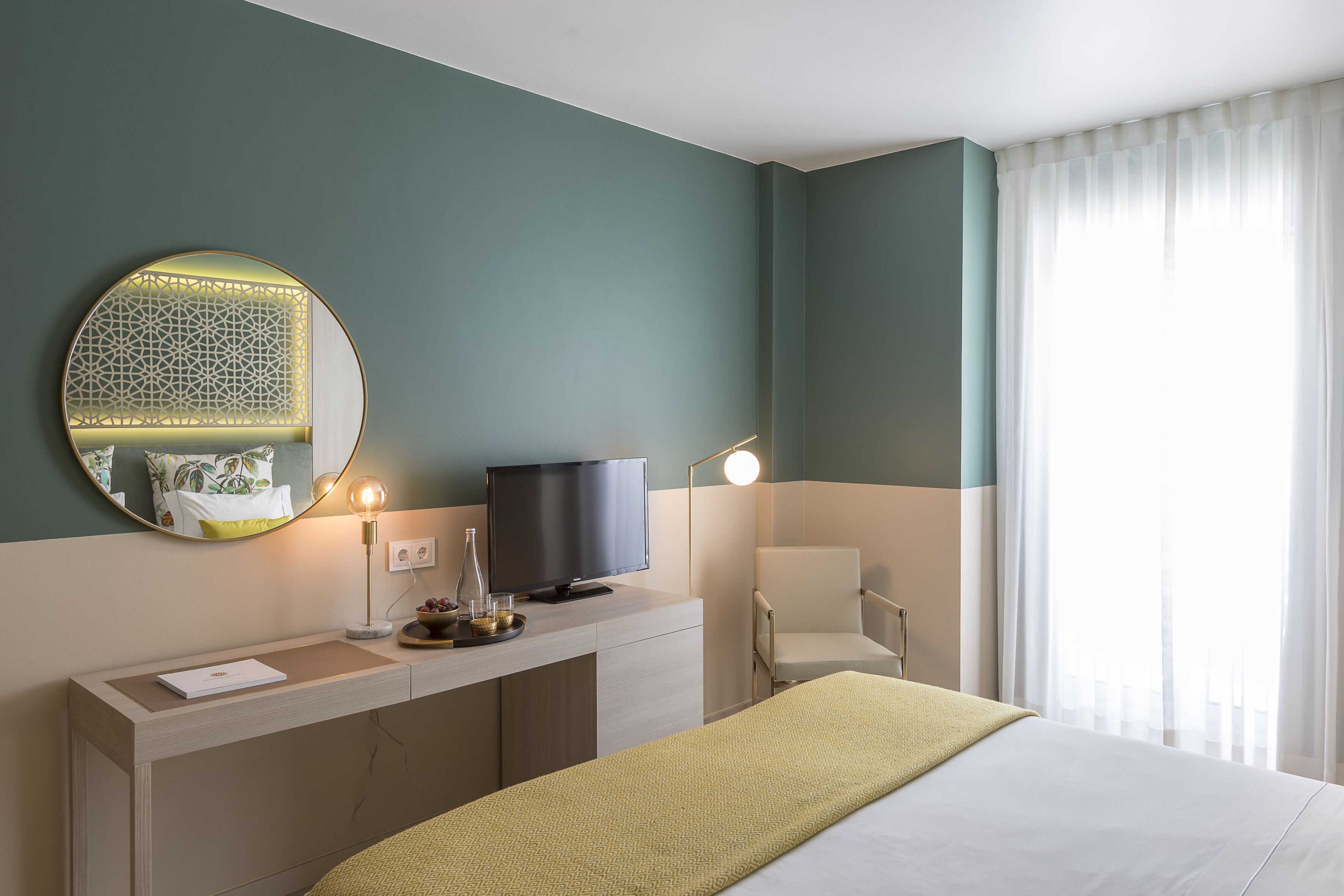 A hotel room with a bed, a desk with a television, a chair, a round mirror, and a window with curtains.