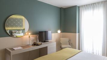 A hotel room with a bed, a desk with a television, a chair, a round mirror, and a window with curtains.