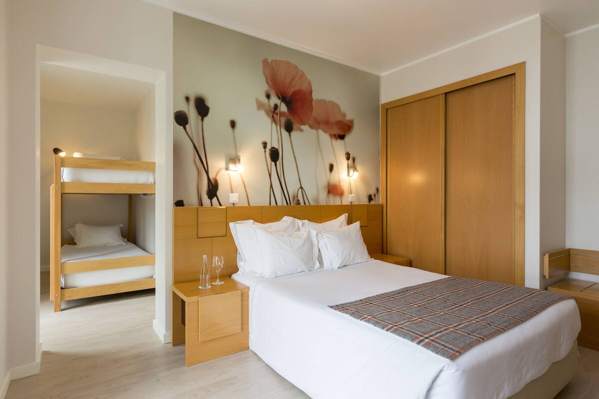 A bedroom with a large bed, a wooden headboard, a nightstand, and a bedside table with a glass. There is a bunk bed in the corner and a wooden wardrobe.