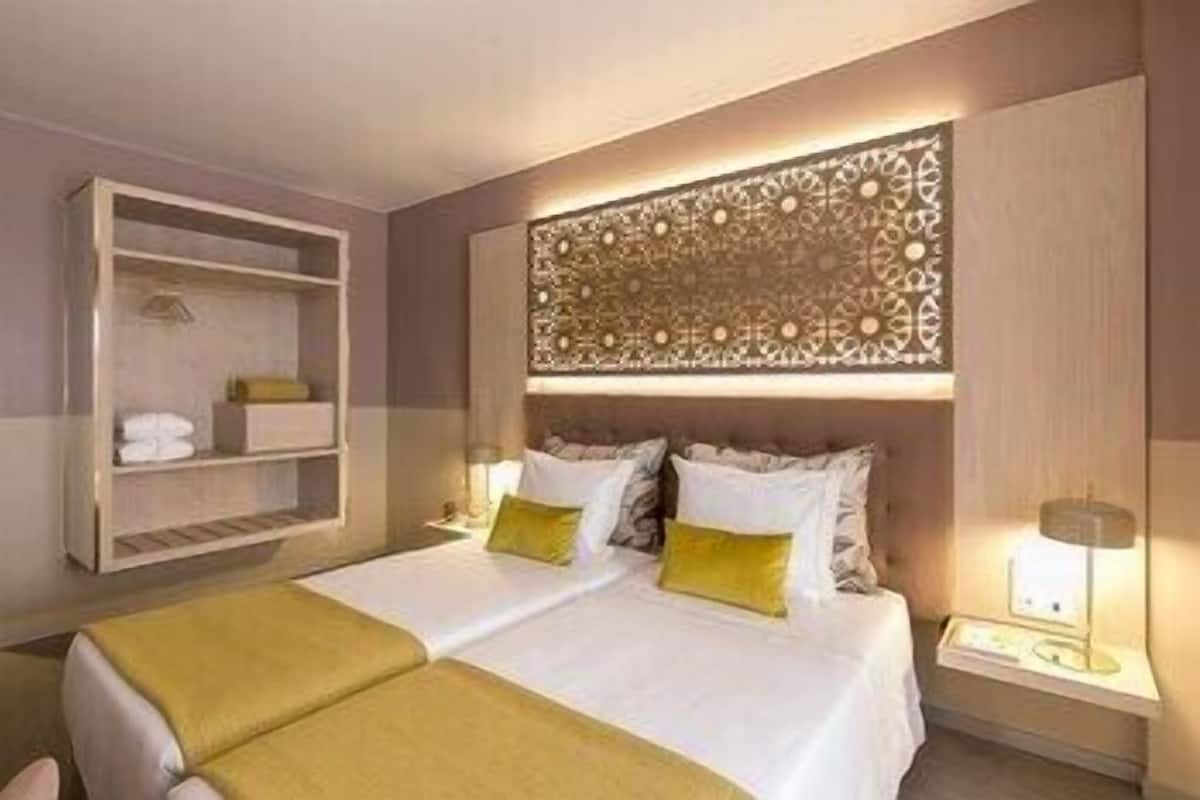 A hotel room with a double bed, two bedside tables, a decorative headboard, and a built-in shelf unit.