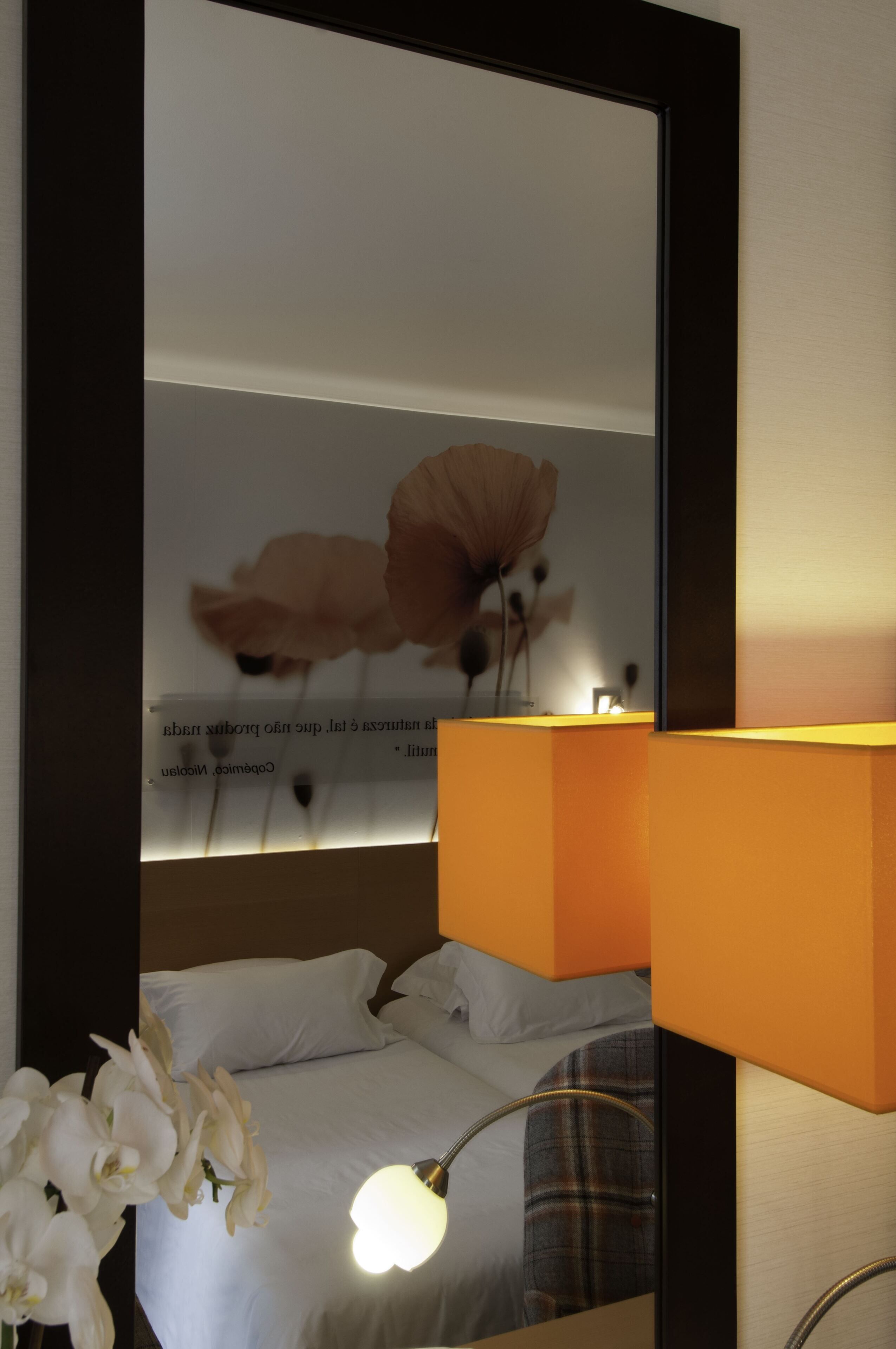 A hotel room with a bed, a nightstand with a lamp, and a wall-mounted mirror reflecting a framed artwork.