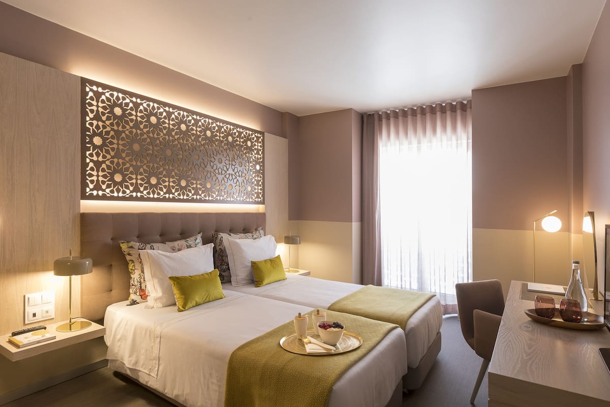 A hotel room with two beds, a desk, a decorative wall panel, and a large window with curtains.