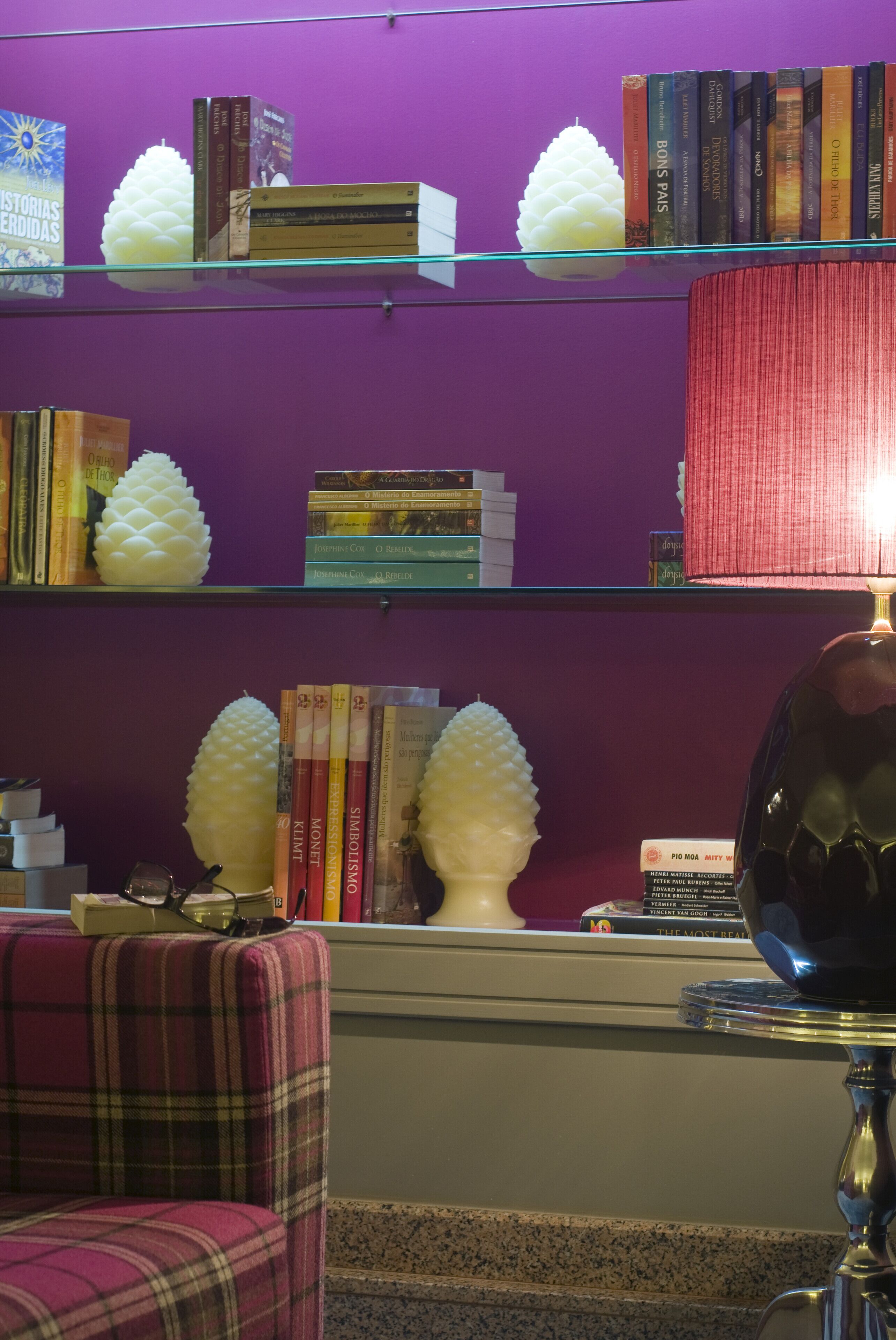 A room with a purple wall, a plaid chair, a table with a lamp, and shelves with books and decorative objects.