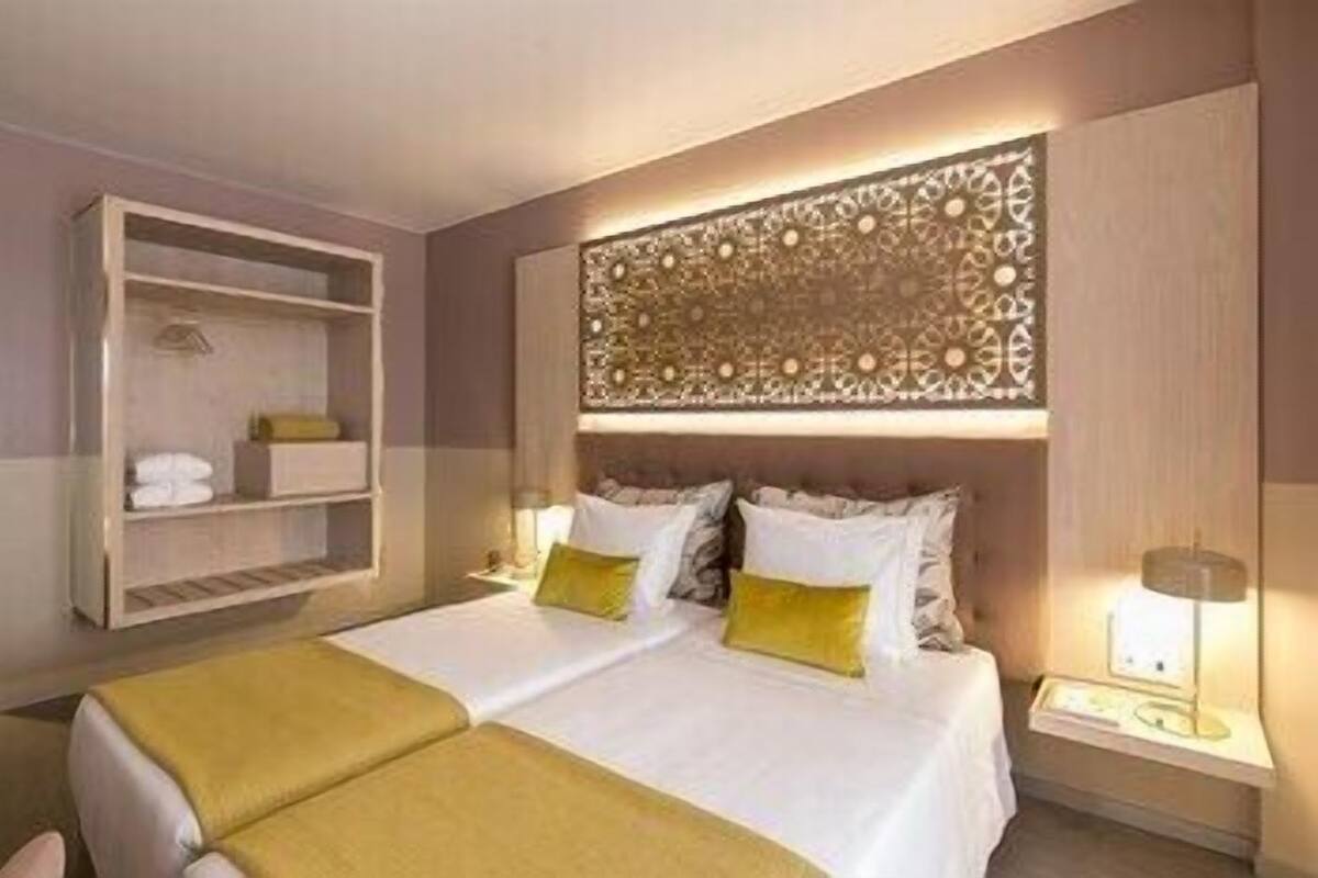 A hotel room with a double bed, two bedside tables, a decorative headboard, and a built-in shelf unit.