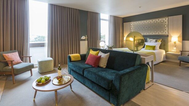A modern hotel room with a green sofa, a dining table with a bottle and glasses, a bed with a headboard, and a large window with curtains. - Hotel Estrela de Fatima (Ourem)